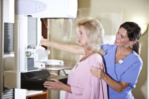 A woman in her 50s getting a mammogram. She is standing at the machine wearing a pink gown. A female technician is atanding behind her, smiling, in blue scrubs, with one hand on the patient's shoulder and smiling.