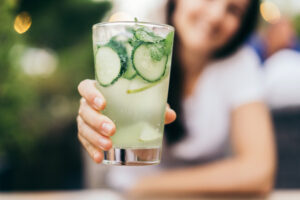 A woman holds out a glass of cucumber lemonade. A cooling non-alcoholic drink on a summer day. Healthy lifestyle.